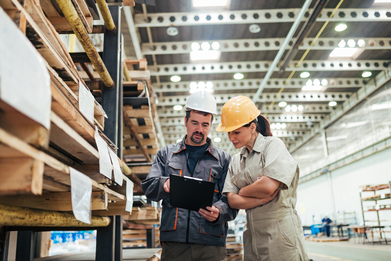 Two people standing next to large shelves in a warehouse, looking at a clipboard