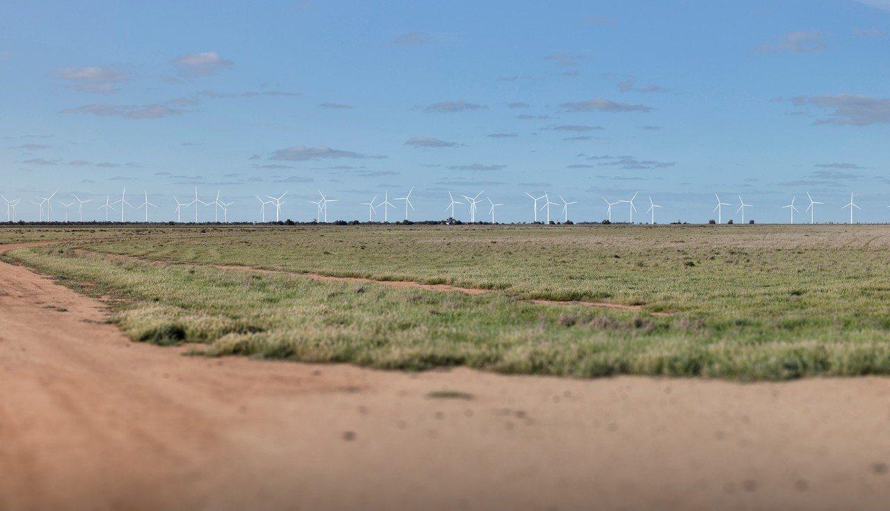An expansive view of Pottinger Energy Park, featuring numerous wind turbines standing tall over the lush green landscape. The turbines' white towers and blades contrast with the natural colors below a partly cloudy sky.