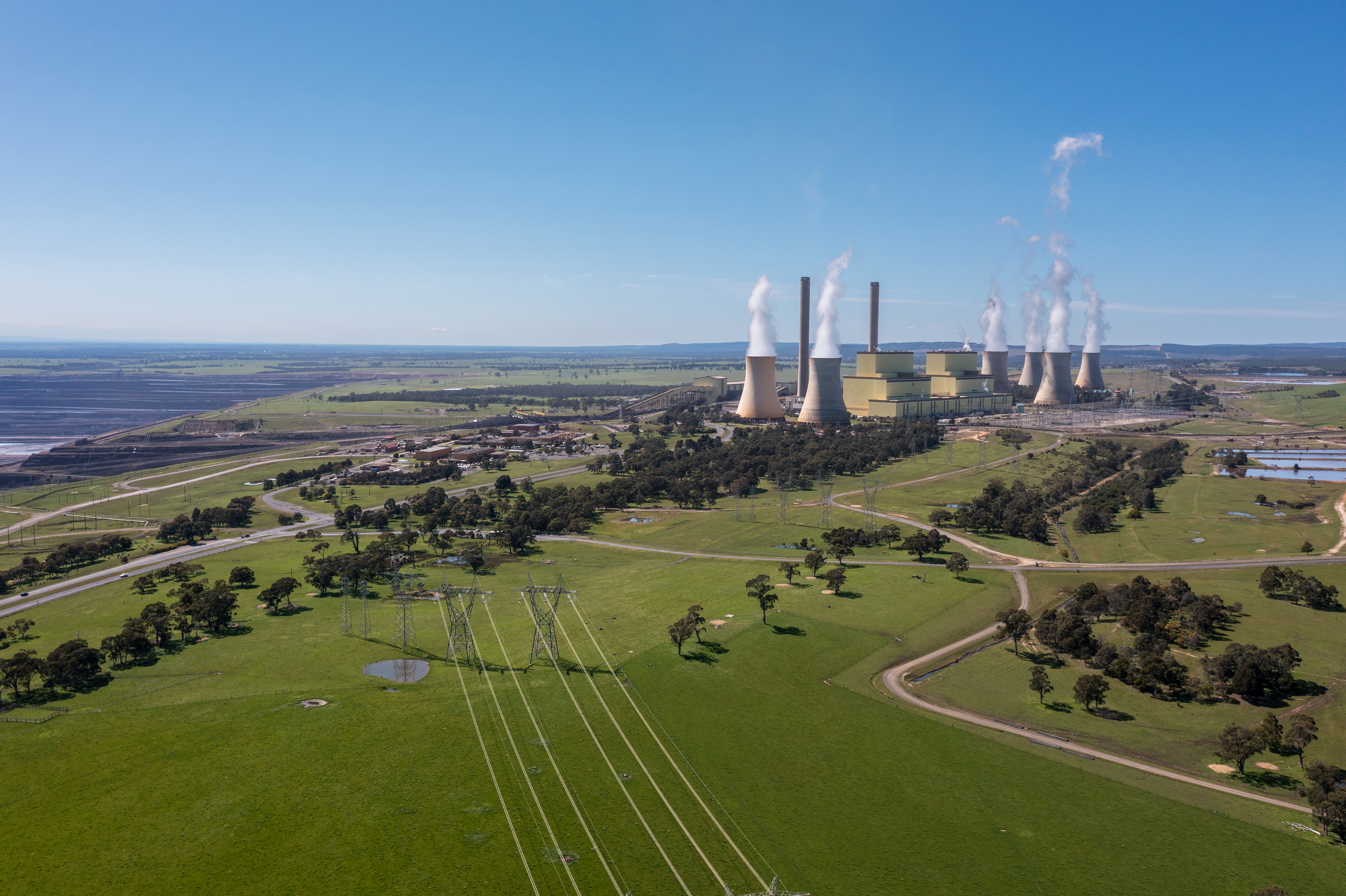 An aerial view of Loy Yang Power Station, with its distinctive cooling towers emitting steam. The facility is set amidst green fields, with a complex network of roads and power lines visible, under a clear blue sky.