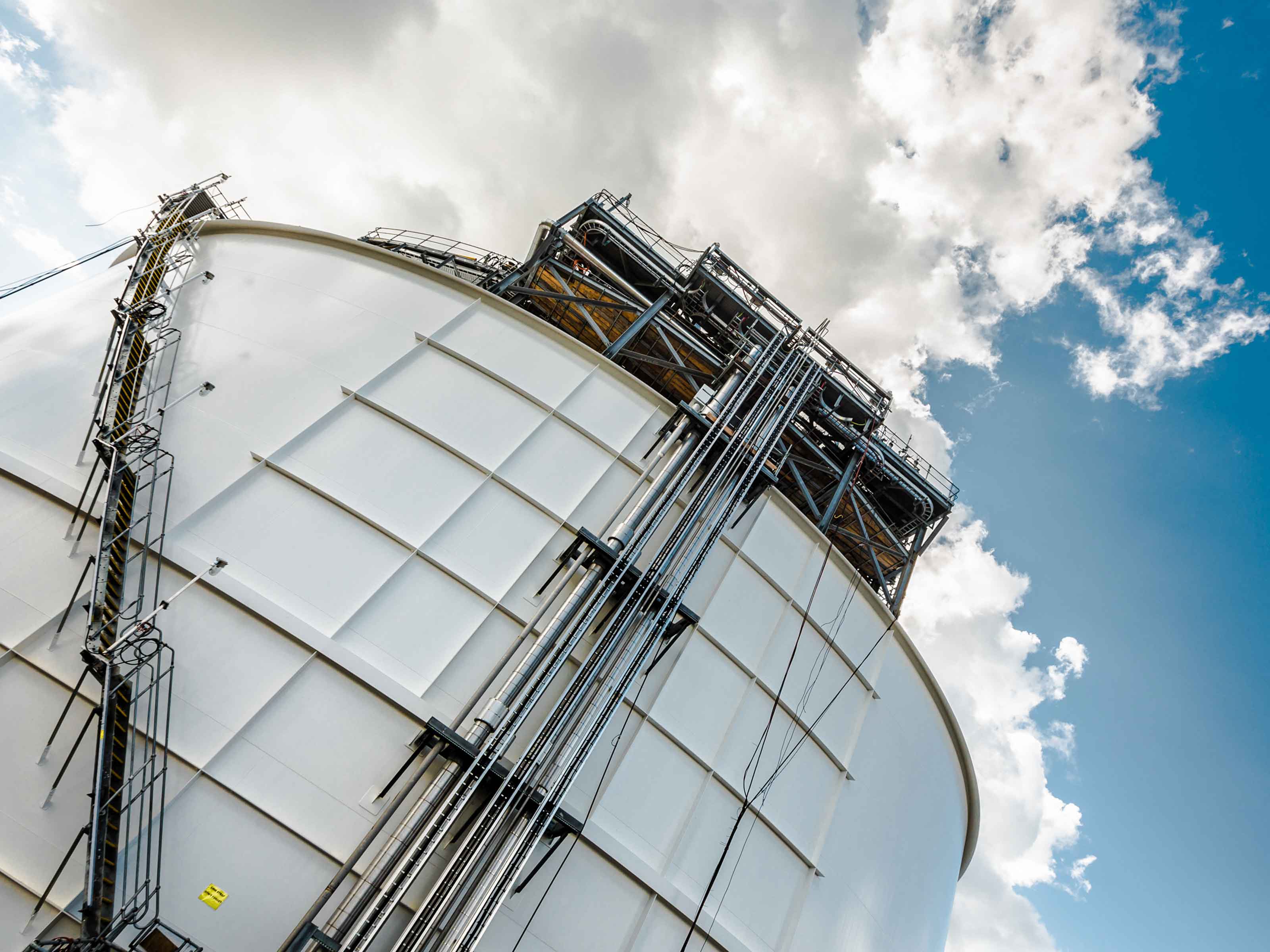 Upward angle view of the Newcastle Gas Storage Facility showing a large white storage tank with intricate piping and scaffolding against a backdrop of blue sky with scattered clouds.