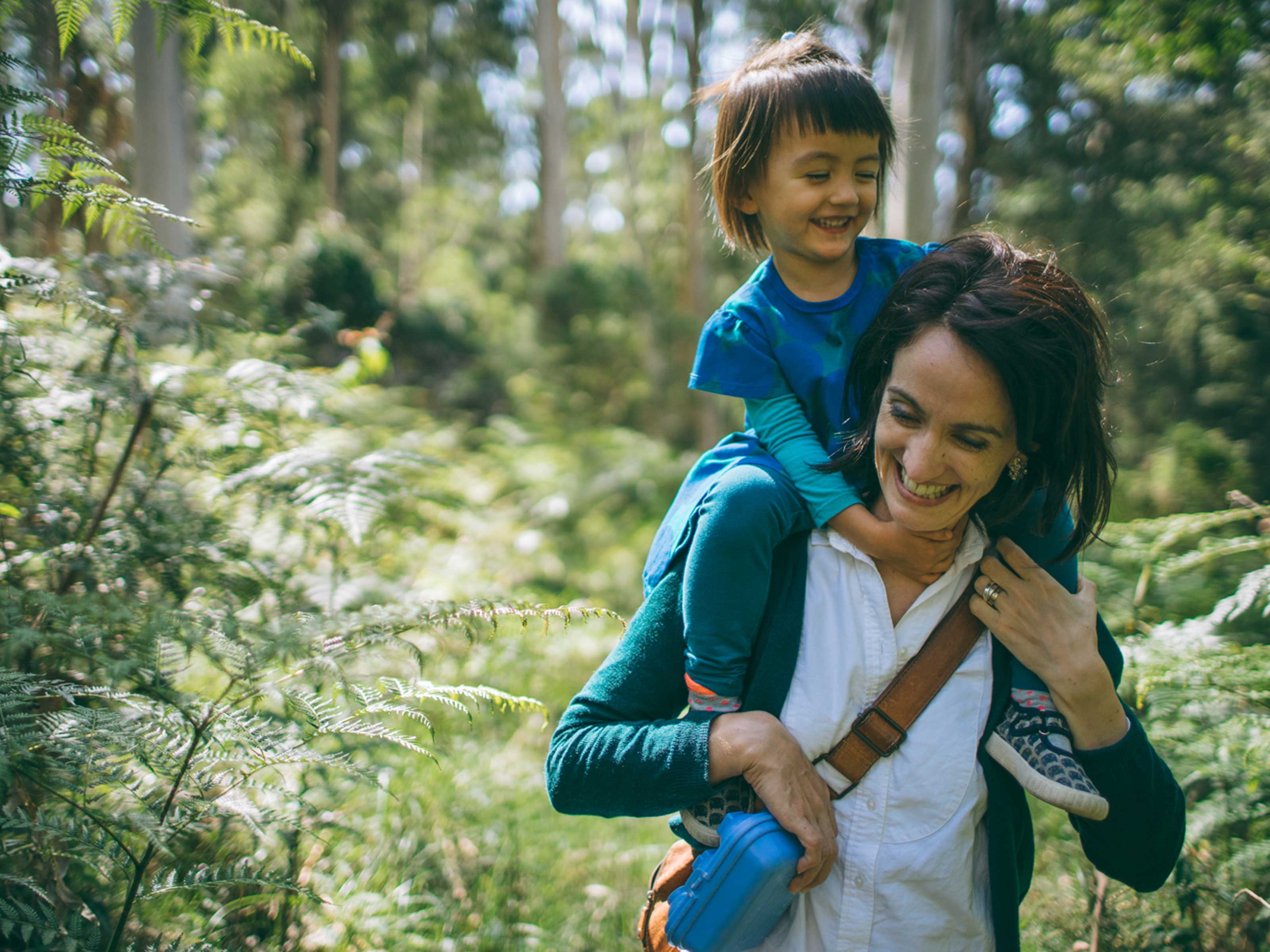 A child on a woman's back laughs in a sunlit forest, evoking a carefree moment in nature.