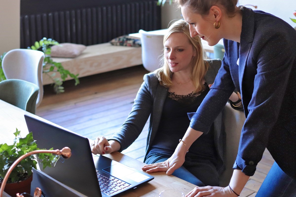Two woman at a desk in an office, on sitting and one standing, looking at a laptop  screen