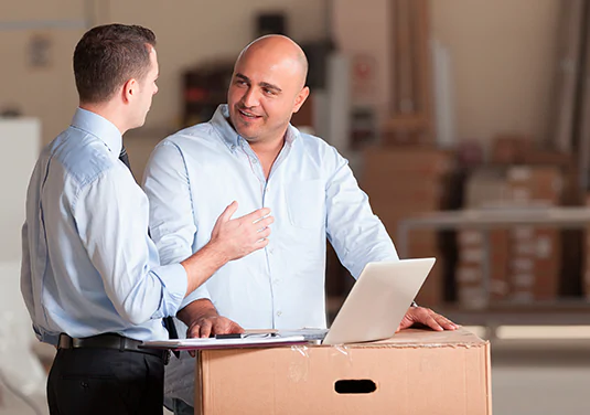 Two men standing in an empty office, discussing something on a laptop that’s resting on a moving box