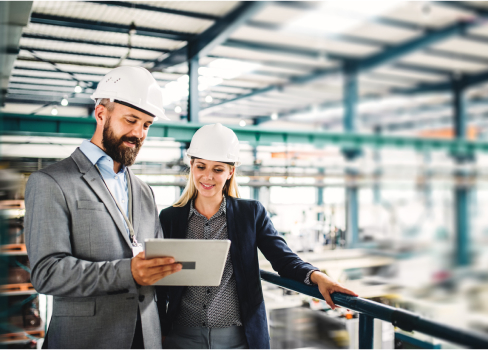 Two coworkers wearing hard hats, smiling and looking at a tablet screen while standing in a warehouse