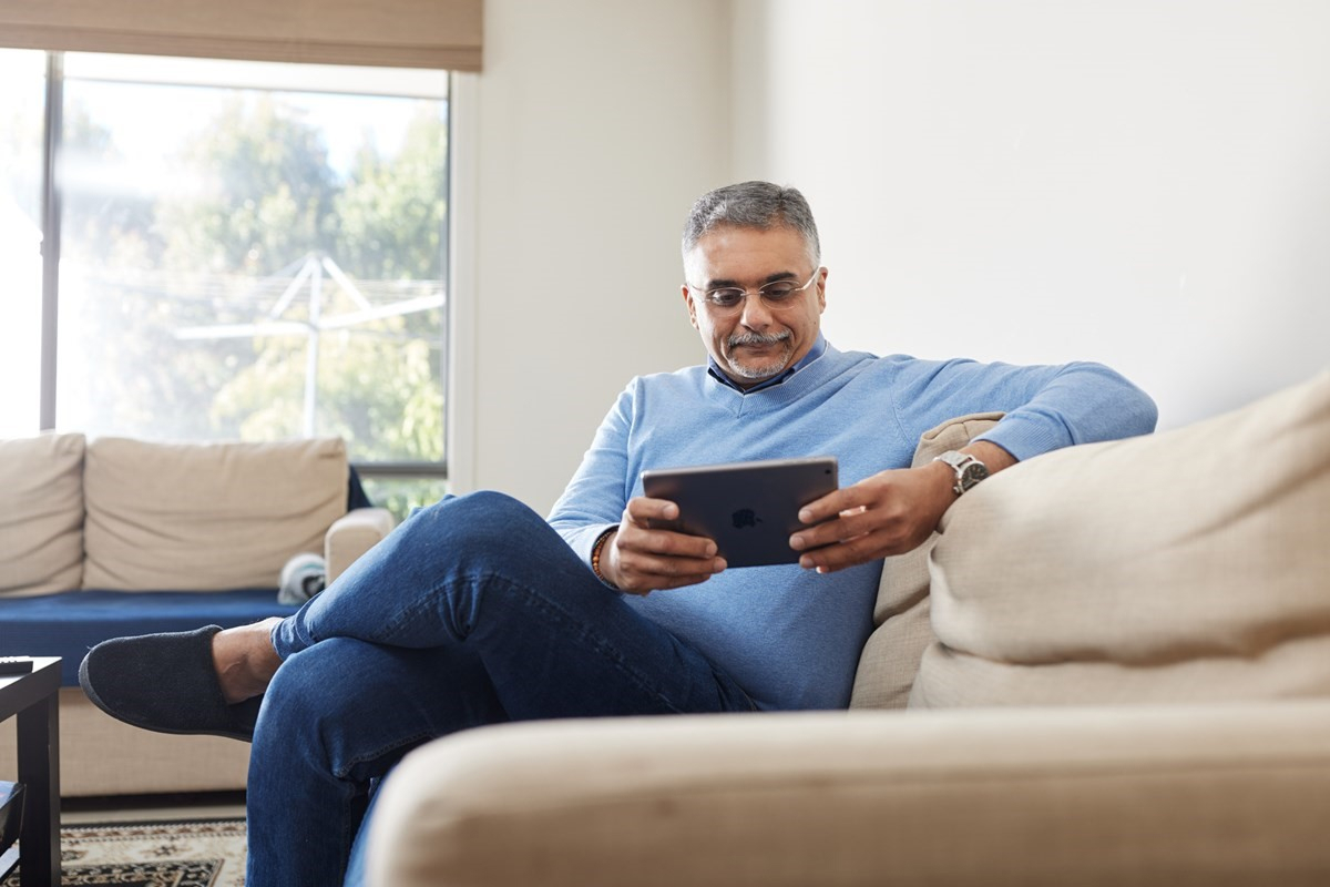 Person in a light blue sweater sitting on a beige couch, looking at a tablet in a sunlit living room.